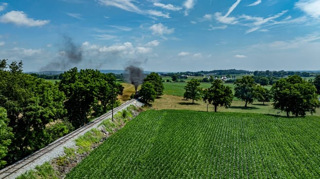 A steam locomotive chugs along tracks surrounded by lush green fields and trees on a bright day. The landscape features a clear blue sky with wispy clouds, enhancing the scenic view.