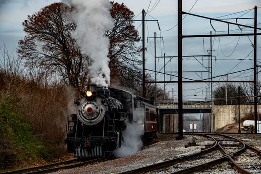 A vintage steam locomotive emits white smoke while pulling a passenger car on railway tracks. The scene is set against a backdrop of trees and an urban area, capturing the essence of railway history.