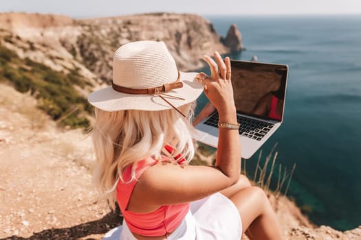 A woman is sitting on a rocky beach with a laptop in front of her. She is wearing a pink top and a straw hat. Concept of relaxation and leisure