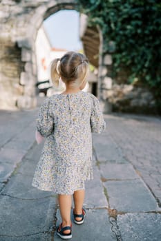 Little girl walks towards an arched passage in a stone wall. Back view. High quality photo