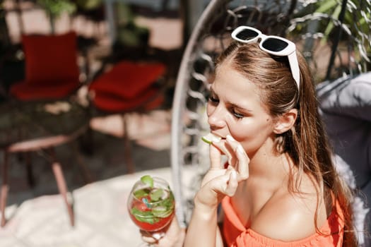 A woman in an orange dress is sitting in a chair with a drink in front of her. She is looking at the camera and she is enjoying her drink. The scene is set in a patio or outdoor area
