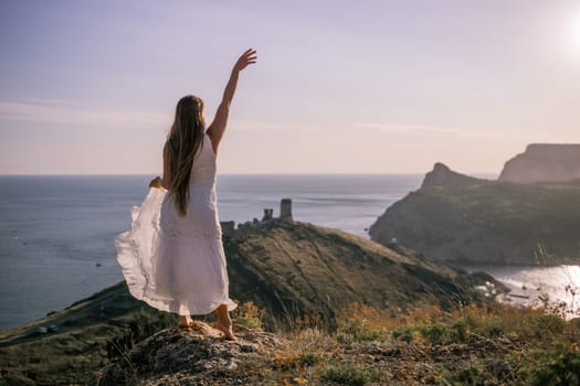 A woman in a white dress stands on a rocky hill overlooking the ocean. She is smiling and she is happy