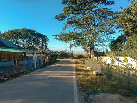 A tranquil rural road stretches between modest homes and lush greenery under a clear blue sky. The serene setting is marred slightly by scattered litter, hinting at human activity amidst the calm.