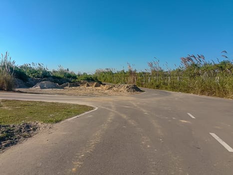 A remote, sunlit road curves through a rural landscape, flanked by tall grasses rustling in the breeze. Mounds of excavated earth and patches of dirt hint at recent construction activity. The clear blue sky evokes tranquility, contrasting with the signs of human disruption.