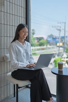 A cheerful businesswoman immersed in her laptop work in a modern office, enjoying a cup of coffee and looking out at an urban view, radiating productivity and focus.
