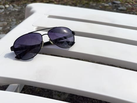 A pair of stylish sunglasses sits on a white lounge chair, surrounded by a peaceful outdoor environment. The sun shines brightly, hinting at a perfect day for relaxation.