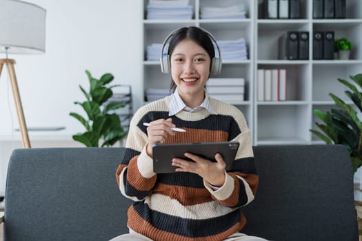 A cheerful young woman enjoys using her tablet in a stylish home office, surrounded by plants and modern decor, highlighting a blend of comfort and creativity.