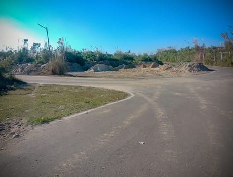 A desolate road curves gently beside a large, barren mound of earth, hinting at recent construction or excavation. Under a clear blue sky, the scene is stark and abandoned, with tall, dry grasses and scattered lamp posts lining the roadside. The mood is one of quiet, forgotten stillness.