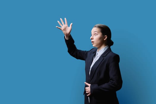 Surprised teenage girl looking in profile at copy space for text of your image, blue studio background. Shocked teenage student in formal wear, education youth emotions