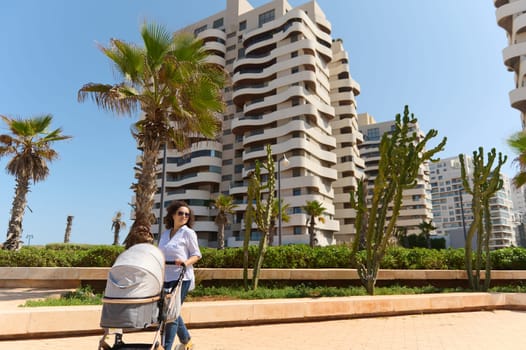 Full length portrait of a happy mother pushing infant baby stroller and walking through the street against high rise buildings and palm trees background. People. Lifestyle. Maternity and babyhood