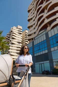 Beautiful multi-ethnic woman, young mom pushing the baby pram, strolling the street in urban environment, standing against modern high rise buildings. People. Parenthood. Babyhood. Maternity lifestyle