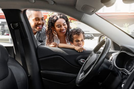 Friendly african american family choosing new car at dealership center parking spot. Excited black mom, dad and preteen child son looking inside new automobile and smiling. Auto buying, transportation