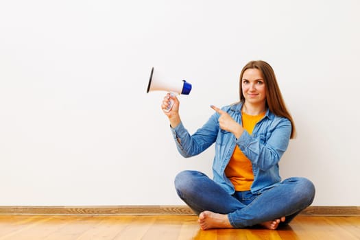 Young beautiful woman with megaphone sitting on wooden floor in front of white wall, copy space.