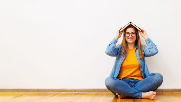 Woman in glasses holding opened book on head while sitting on the floor near white wall with copy space. Concept of education, organization of courses, conducting training classes