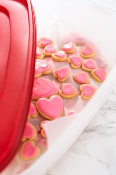 Storing heart-shaped sugar cookies with pink and white royal icing in a large plastic container.