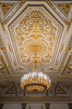 An ornate hall features a stunning chandelier beneath a beautifully detailed ceiling, showcasing elaborate gold leaf patterns and classical architectural design elements.
