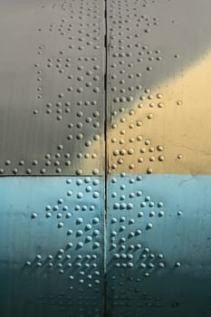 Close-up view of a textured industrial surface featuring colorful sections and rivets. The image highlights the interplay of light, shadow, and color on the metal.
