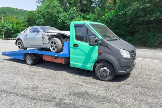 A tow truck carries a silver car with damage on a blue flatbed along a rural road. Lush greenery surrounds the area, indicating a sunny day.