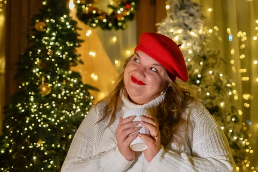A woman in a red beret and white sweater holds a mug and looks up at Christmas lights.