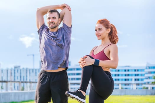 man and woman stretch their muscles during a workout session in an urban park. The man raises his arms while the woman lifts her knee, both dressed in sporty attire and enjoying a sunny, active day.