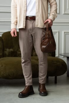 A fashionable man stands with a brown hip bag in a contemporary room, embodying urban style and professionalism. Beige shirt and brown pants
