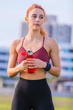 Fit young woman in sporty attire holds a red water bottle while standing outdoors. She looks off to the side with a focused expression, surrounded by an urban park setting on a sunny day