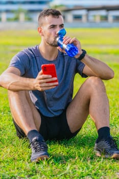 A fit man sits on the grass in a park, drinking water from a blue bottle while holding a red smartphone. He wears sporty attire, resting after exercise on a sunny day in a natural outdoor setting.