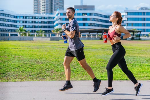 Fit man and woman jogging together in a sunny park, holding water bottles. Both dressed in sporty attire, smiling and enjoying outdoor exercise surrounded by green grass and trees