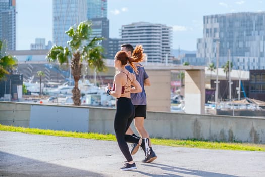 Fit man and woman jogging together in a sunny park, holding water bottles. Both dressed in sporty attire, smiling and enjoying outdoor exercise surrounded by green grass and trees