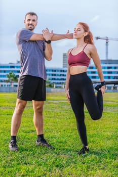 man and woman stretch their muscles during a workout session in an urban park. The man raises his arms while the woman lifts her knee, both dressed in sporty attire and enjoying a sunny, active day.