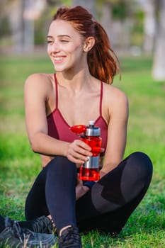 A fit young woman with red hair sits on the grass holding a red water bottle. She is dressed in sporty attire, smiling and enjoying a sunny day outdoors in a natural park setting after exercising.