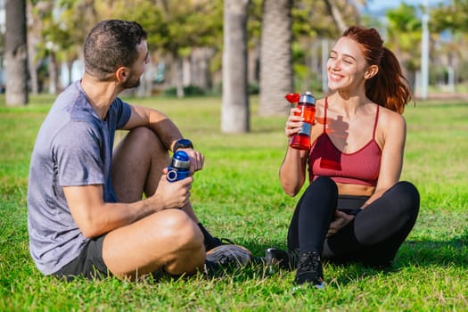 A fit man and woman sit on the grass in a sunny park, smiling and holding water bottles. They are dressed in sporty attire, relaxing and staying hydrated after a workout session surrounded by trees and nature