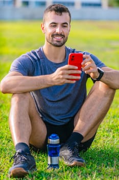 Man Young athletic man sitting cross-legged on green grass in a park, holding a red smartphone. He appears relaxed, smiling, and dressed in sporty clothes with a water bottle nearby. Vertical