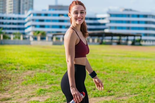 A fit young woman in sporty attire smiles while holding a red water bottle. She stands in an urban park with green grass and modern buildings in the background, enjoying a sunny outdoor workout.
