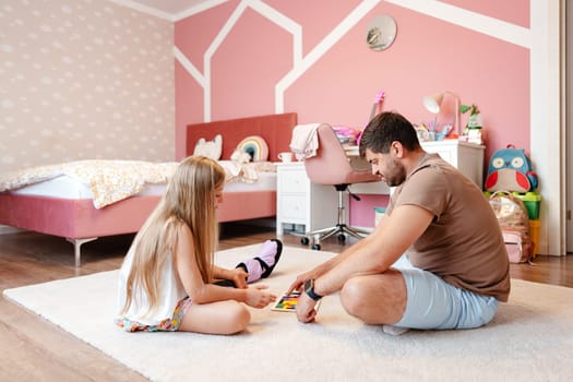 A young girl is playing a game with her father on the floor in a bedroom. The room is decorated in pink and white with a bed in the background.