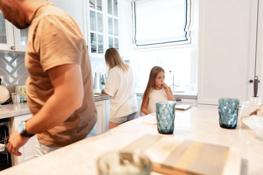 A family is in their kitchen. The father is loading the dishwasher, while the mother is washing dishes. The daughter is sitting at the counter eating a snack.