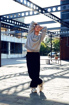 A man wearing a gray sweatshirt, black pants, and white sneakers stands with his arms raised above his head. He is in a paved courtyard with a metal structure overhead.