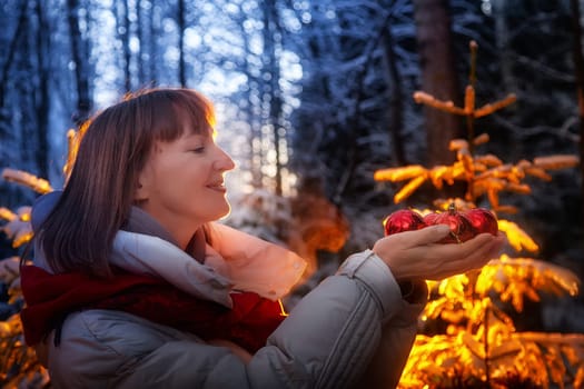 A middle-aged woman smiles as she holds red ornaments in her hands while exploring a snowy forest, surrounded by Christmas spirit.
