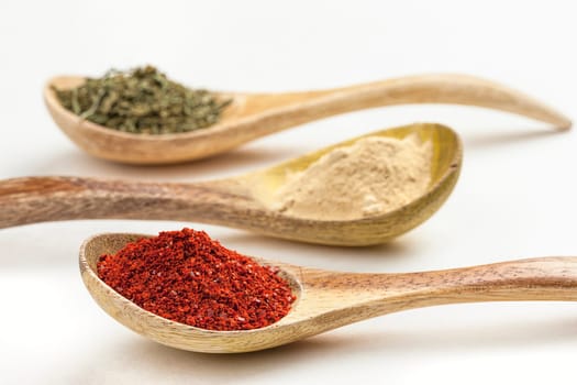 A close up of wooden spoons filled with red pepper powder, garlic powder, and basil leaves.