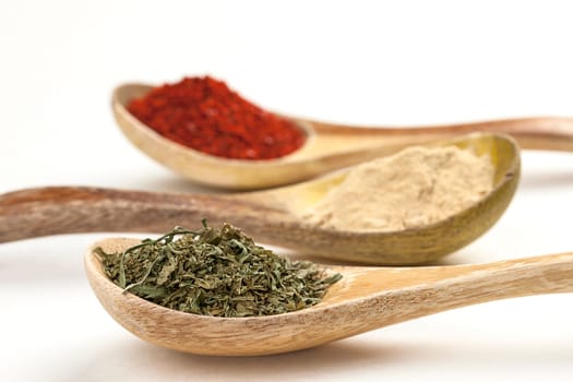 A close up of wooden spoons filled with red pepper powder, garlic powder, and basil leaves.