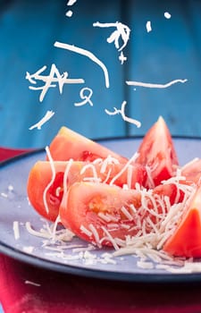 Shredded white cheese being sprinkled onto tomato wedges on a plate.