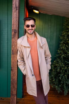 Confident handsome man in beige trench coat standing with hands in pockets outside green house. Young casual good looking male is leaning on wooden pole outdoors during Christmas.