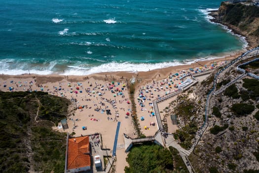 Aerial drone view of unrecognizable tourists on sandy seashore at Magoito Beach. Distant shot of sea waves splashing on coast of famous destination in Sintra, Portugal during summer.