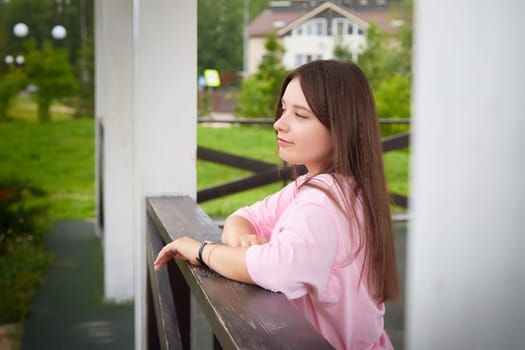 A young woman with long brown hair is wearing a pink shirt and is leaning against a white post