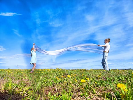 A child and an adult happily wave colorful fabric in a vibrant green field filled with wildflowers under a clear blue sky.