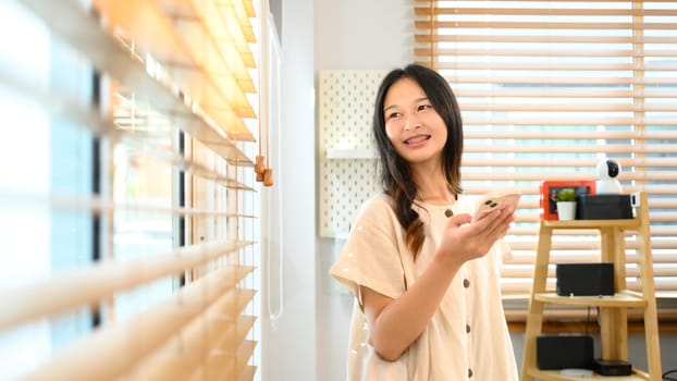 Pretty young woman holding smartphone and looking out window at home.