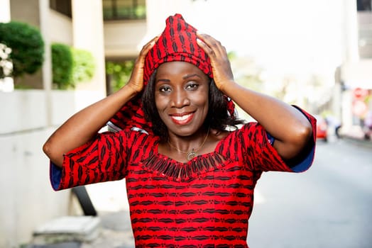mature african woman standing outdoors adjusting her scarf and looking at the camera smiling.