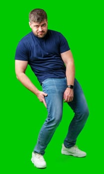 A man in a blue shirt and jeans awkwardly poses against a bright green backdrop in studio
