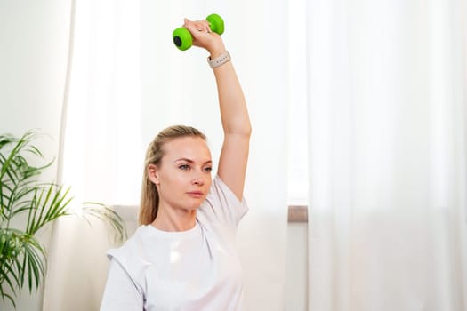 A woman exercises with green dumbbells at home in a bright room filled with light and plants