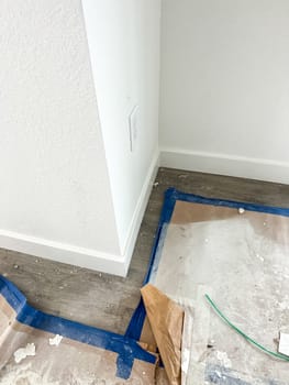 Parker, Colorado, USA-January 11, 2023-A close-up view of a corner in a new construction home featuring newly installed baseboards, a finished white wall, and flooring partially covered with protective paper and painter tape. Construction debris and materials are visible, indicating ongoing work.